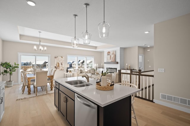 kitchen featuring a raised ceiling, a kitchen island with sink, stainless steel dishwasher, and decorative light fixtures