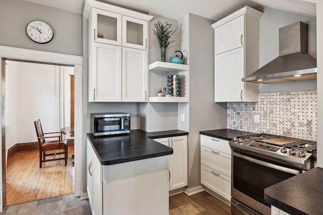 kitchen featuring dark countertops, stainless steel appliances, white cabinets, wall chimney exhaust hood, and glass insert cabinets