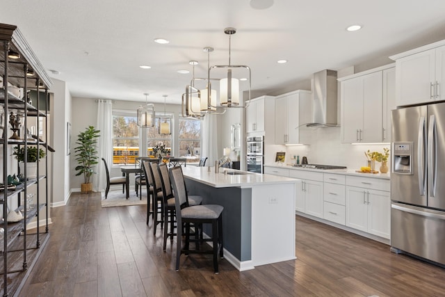 kitchen featuring stainless steel appliances, an island with sink, wall chimney range hood, and dark wood-style flooring