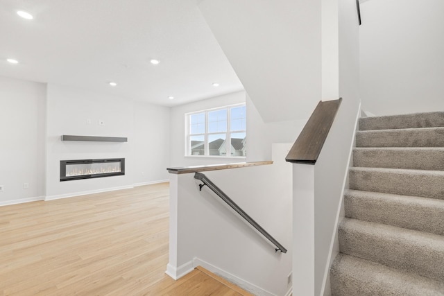 staircase featuring baseboards, a glass covered fireplace, wood finished floors, and recessed lighting