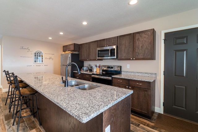 kitchen with dark wood-style floors, a center island with sink, appliances with stainless steel finishes, a sink, and dark brown cabinets