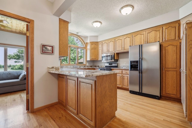 kitchen with appliances with stainless steel finishes, kitchen peninsula, light stone countertops, a textured ceiling, and light hardwood / wood-style flooring