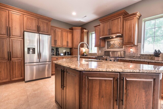 kitchen featuring sink, stainless steel fridge, a kitchen island with sink, light stone counters, and decorative backsplash