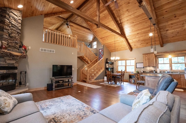 living room with a stone fireplace, dark hardwood / wood-style floors, ceiling fan with notable chandelier, beamed ceiling, and wooden ceiling