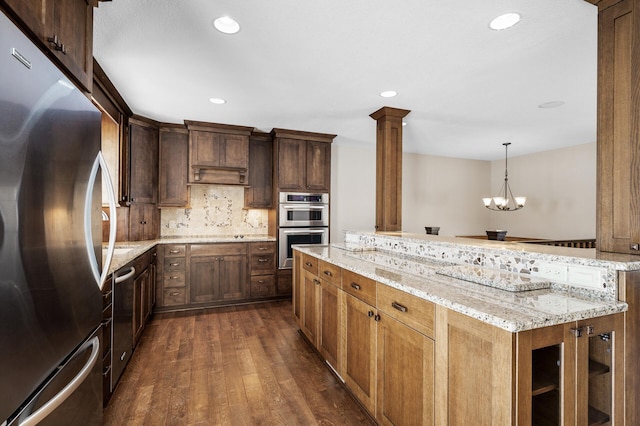 kitchen with light stone counters, stainless steel appliances, dark wood-style flooring, decorative columns, and pendant lighting