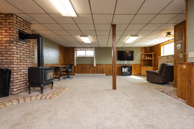 interior space featuring a paneled ceiling, light carpet, a wood stove, baseboard heating, and wooden walls