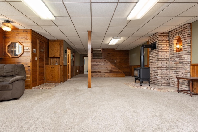 basement with carpet floors, a wood stove, a paneled ceiling, and wooden walls