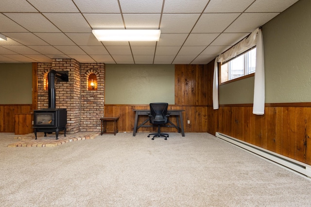 carpeted office featuring a baseboard radiator, a drop ceiling, a wood stove, and wood walls