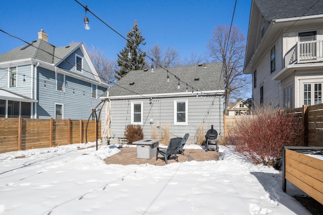 snow covered house with an outdoor fire pit, a shingled roof, and a fenced backyard