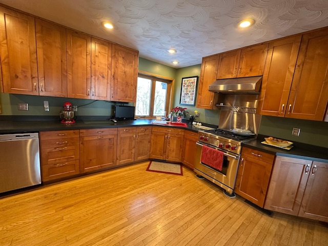 kitchen with light wood-type flooring, dark countertops, brown cabinetry, under cabinet range hood, and appliances with stainless steel finishes