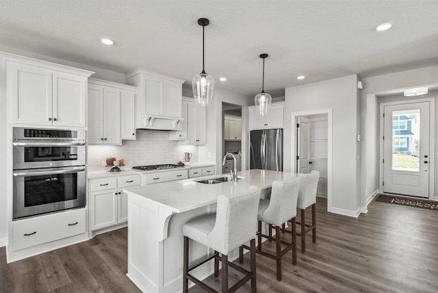 kitchen featuring sink, appliances with stainless steel finishes, white cabinetry, hanging light fixtures, and an island with sink