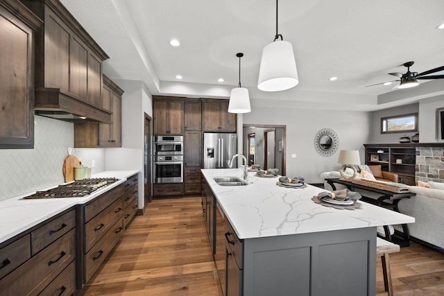 kitchen with pendant lighting, sink, light stone counters, stainless steel appliances, and a center island with sink