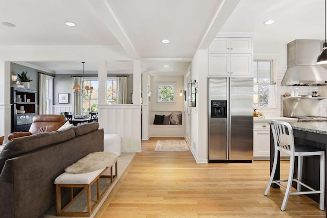 kitchen featuring light wood-type flooring, stainless steel refrigerator with ice dispenser, white cabinetry, wall chimney range hood, and decorative backsplash