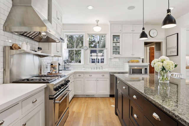 kitchen with light wood-type flooring, decorative light fixtures, white cabinetry, stainless steel appliances, and wall chimney exhaust hood