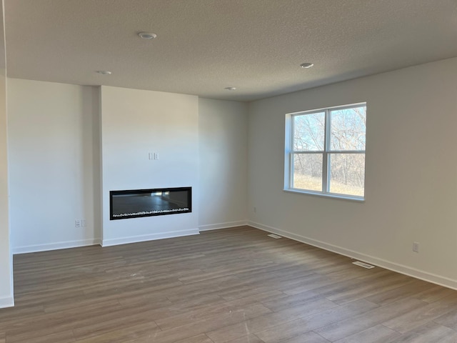 unfurnished living room with a textured ceiling, visible vents, baseboards, light wood-style floors, and a glass covered fireplace