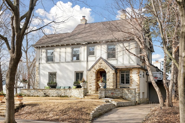 english style home with a shingled roof, a chimney, driveway, and stucco siding