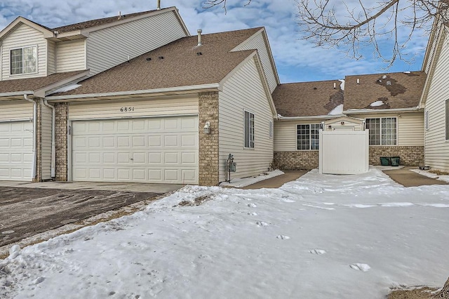 view of snowy exterior with an attached garage, driveway, roof with shingles, and brick siding