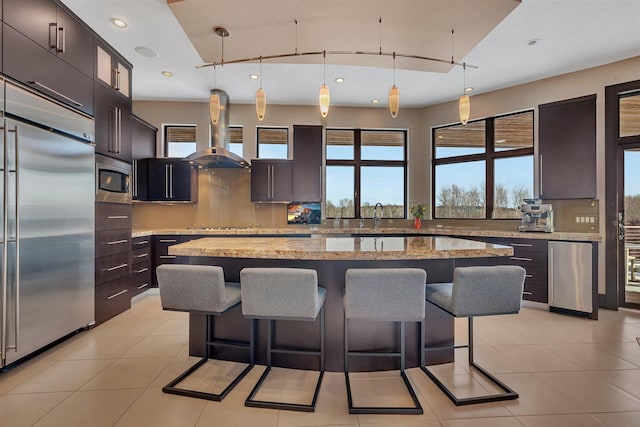 kitchen featuring dark brown cabinetry, light stone counters, built in appliances, a center island, and pendant lighting