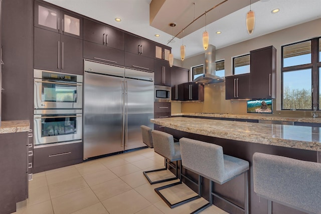 kitchen with light stone counters, built in appliances, hanging light fixtures, dark brown cabinets, and island exhaust hood