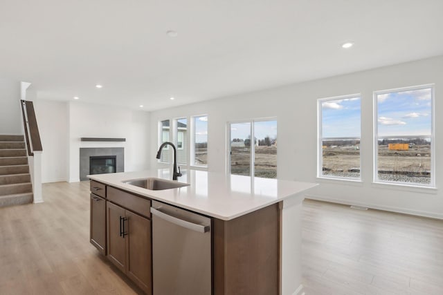 kitchen with sink, a tiled fireplace, a kitchen island with sink, stainless steel dishwasher, and light hardwood / wood-style floors