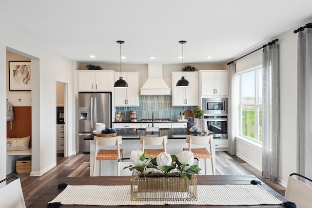 kitchen featuring premium range hood, appliances with stainless steel finishes, tasteful backsplash, an island with sink, and hanging light fixtures