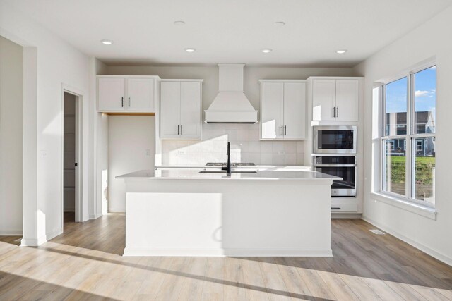 kitchen featuring premium range hood, a center island with sink, light wood-type flooring, stainless steel appliances, and white cabinets