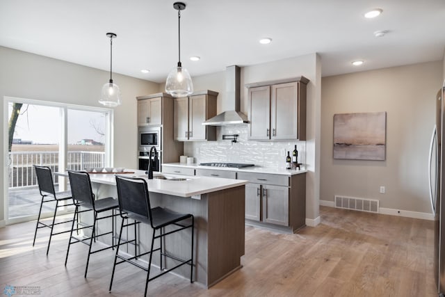 kitchen with sink, pendant lighting, stainless steel appliances, a kitchen island with sink, and wall chimney range hood