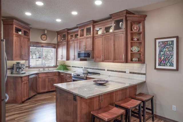 kitchen featuring brown cabinetry, wood finished floors, a peninsula, a sink, and appliances with stainless steel finishes