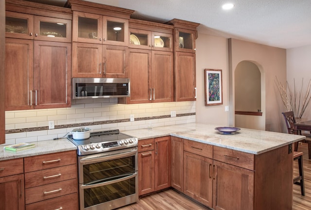 kitchen featuring light wood-style flooring, stainless steel appliances, a peninsula, brown cabinetry, and decorative backsplash