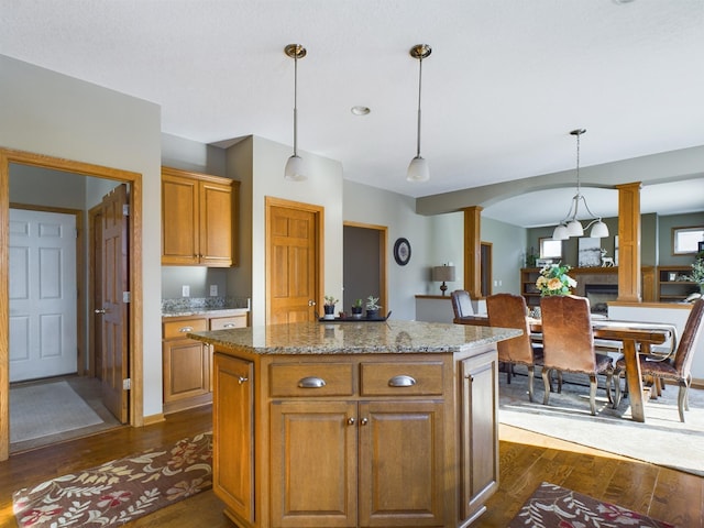 kitchen featuring light stone counters, a kitchen island, hanging light fixtures, brown cabinetry, and decorative columns