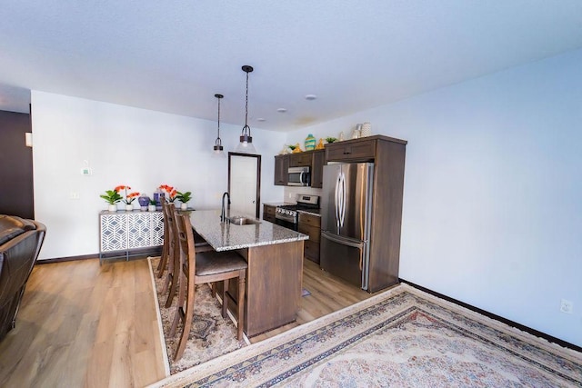 kitchen with sink, appliances with stainless steel finishes, dark brown cabinetry, light stone countertops, and an island with sink