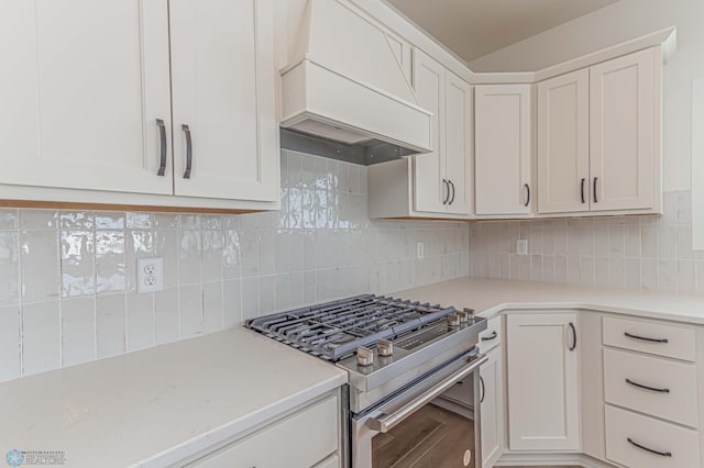 kitchen with custom exhaust hood, stainless steel gas range oven, white cabinetry, light stone counters, and backsplash
