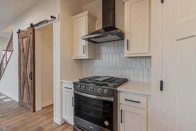 kitchen featuring wall chimney range hood, stainless steel range with gas stovetop, a barn door, light countertops, and light wood-style flooring