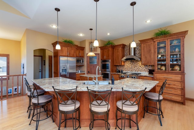 kitchen with appliances with stainless steel finishes, a large island with sink, wall chimney range hood, a kitchen bar, and pendant lighting