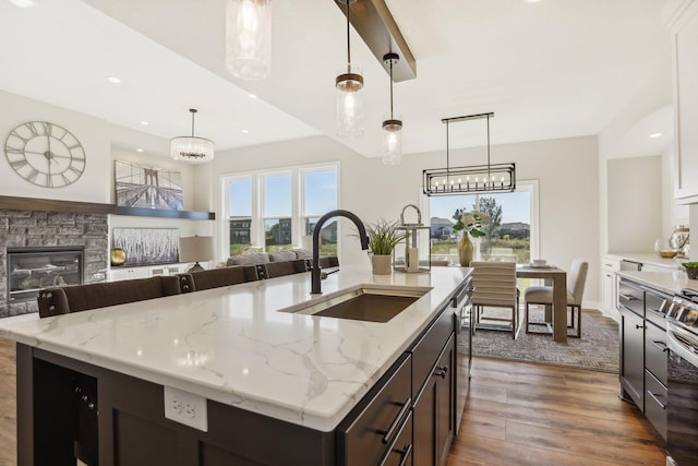 kitchen with a stone fireplace, pendant lighting, an island with sink, sink, and dark hardwood / wood-style flooring