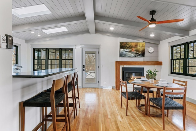 dining area featuring visible vents, baseboards, a glass covered fireplace, light wood-style flooring, and vaulted ceiling with beams