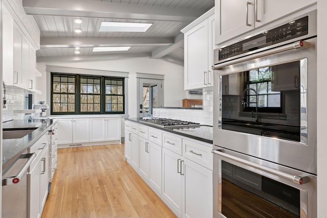 kitchen featuring light wood finished floors, stainless steel appliances, tasteful backsplash, lofted ceiling with skylight, and white cabinetry