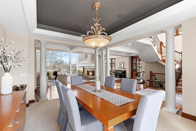 dining room with light colored carpet, stairway, a tray ceiling, a glass covered fireplace, and ornate columns