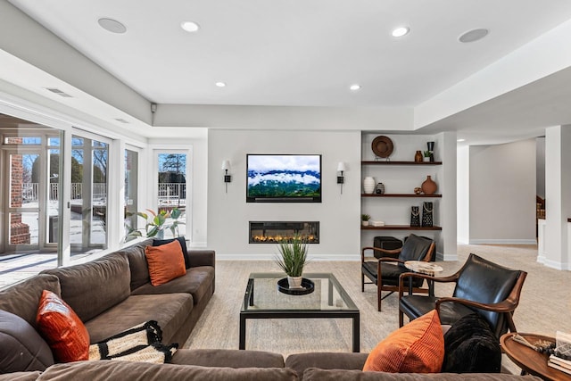 living room featuring built in shelves, recessed lighting, a glass covered fireplace, light carpet, and baseboards