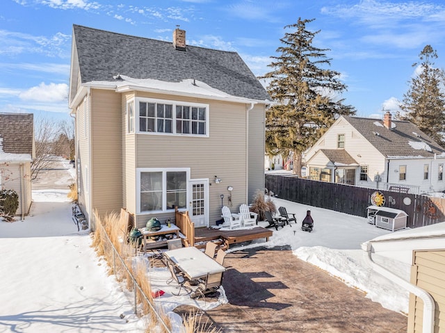 snow covered property with a shingled roof, a chimney, fence, and a deck