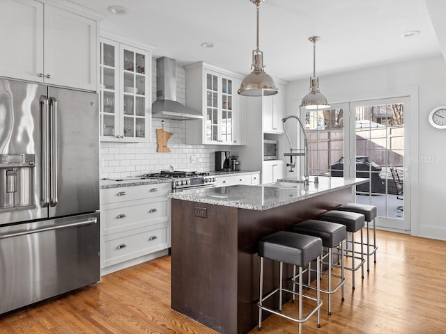 kitchen featuring high end appliances, wall chimney range hood, a kitchen island with sink, and white cabinets