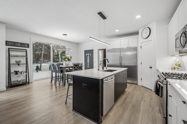 kitchen featuring pendant lighting, appliances with stainless steel finishes, white cabinetry, an island with sink, and light wood-type flooring