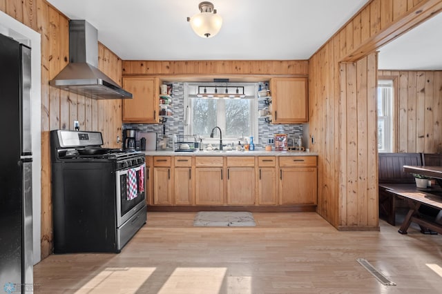 kitchen featuring wooden walls, stainless steel appliances, a sink, light countertops, and wall chimney range hood