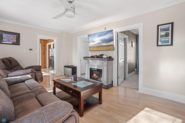 living room with ornamental molding, a glass covered fireplace, ceiling fan, light wood-type flooring, and baseboards