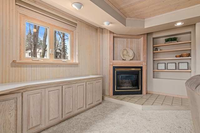 living area featuring ornamental molding, a glass covered fireplace, light colored carpet, built in shelves, and wooden ceiling