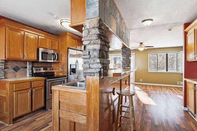 kitchen with dark wood-type flooring, ceiling fan, a kitchen bar, appliances with stainless steel finishes, and ornate columns