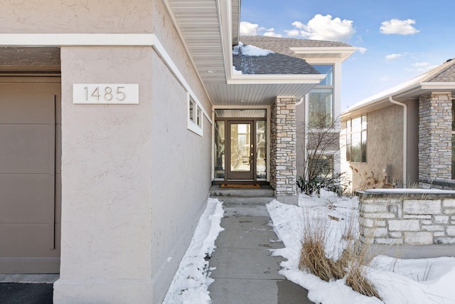 snow covered property entrance with stucco siding, stone siding, and a shingled roof