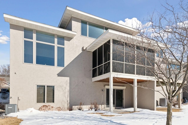 snow covered back of property featuring central air condition unit, a patio area, a sunroom, and stucco siding