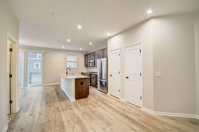 kitchen with appliances with stainless steel finishes, sink, a kitchen island with sink, dark brown cabinets, and light hardwood / wood-style flooring