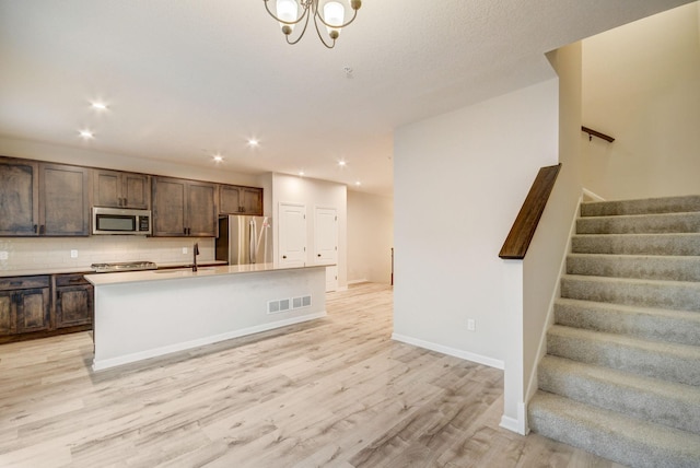 kitchen with tasteful backsplash, stainless steel appliances, dark brown cabinets, a center island with sink, and light hardwood / wood-style flooring